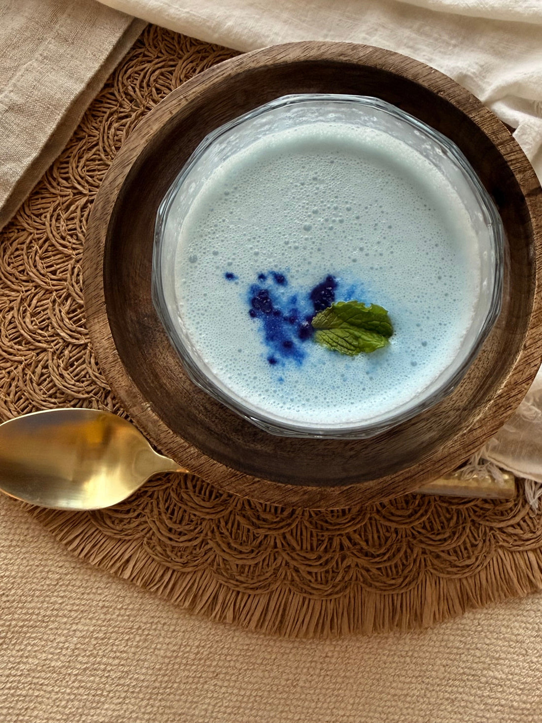 Overhead view of a pale-blue coconut smoothie made with blue spirulina, banana, Greek yogurt, and mint, styled with natural linens, warm wood, and a gold spoon.